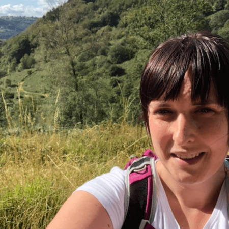 Jo is wearing a white t-shirt and carrying a rucksack. She has dark hair and a fringe. In the background are grasses and trees.